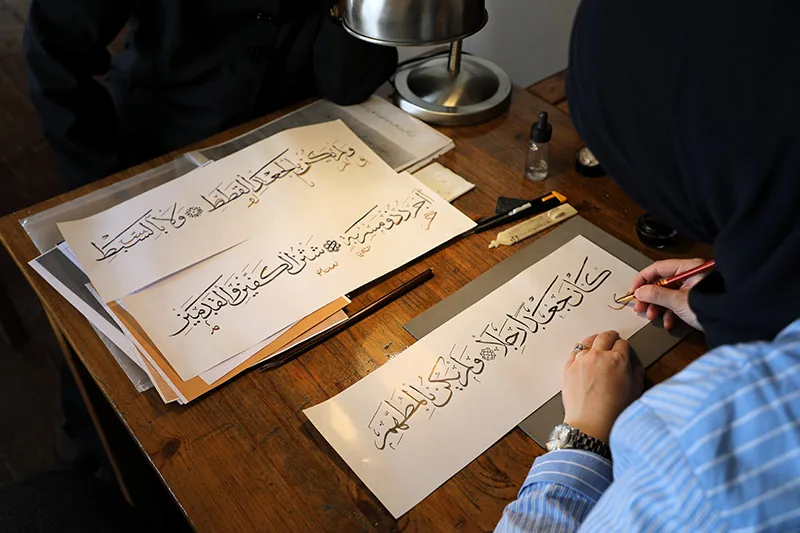 Iemand schrijft met een rietpen grote Arabische letters aan een werktafel