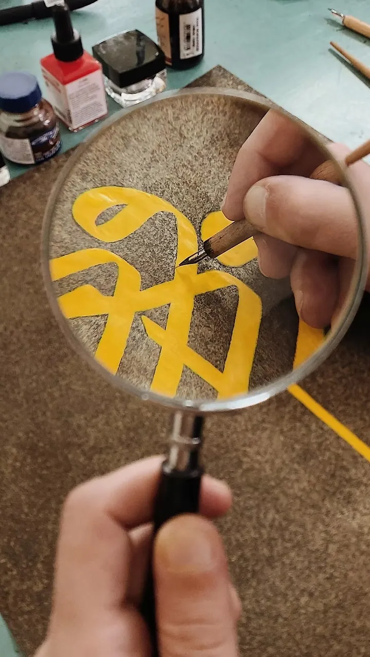 Een gele letter wordt door een vergrootglas bekeken en met een fijne pen bijgewerkt